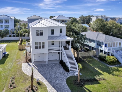 Modern elevated beach house with metal roof and covered parking, nestled among palm trees in coastal neighborhood.