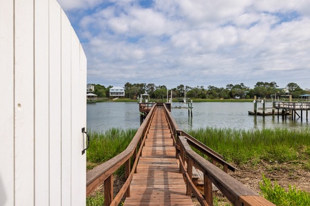 A private dock stretches across calm waters toward neighboring homes and boat slips, framed by marshland grasses and distant trees under coastal skies.