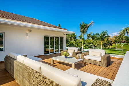 Relax on the outdoor patio with wicker-style seating, offering comfortable white cushions and a serene view of the lush surroundings.