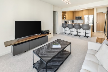 Bright living area with a large TV, modern furnishings, and a view into the kitchen.