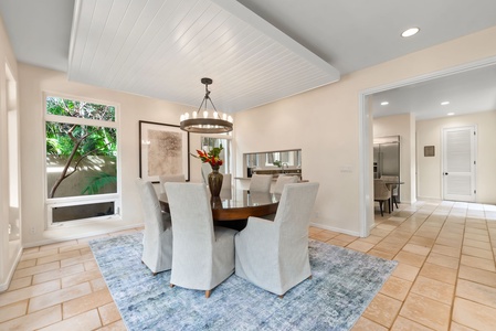Your elegant dining space features a stunning round table for eight beneath a dramatic chandelier, with tropical garden views through expansive windows.