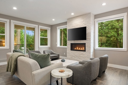 Unwind in this elegant living space with a cozy fireplace, plush seating, and peaceful forest views through expansive windows.