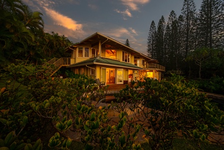 Tropical vacation home glowing warmly at sunset, surrounded by lush palms and towering pines in a serene island setting.