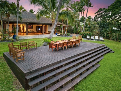 Tropical villa surrounded by lush palm trees and landscaped gardens, featuring an elevated wooden deck with outdoor dining space beneath a colorful sunset sky.
