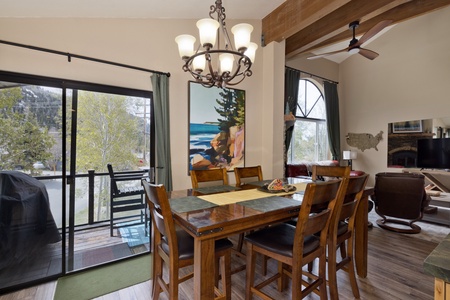 Picture yourself gathered around this handsome wooden dining table, where natural light pours through tall windows and exposed beams add rustic mountain charm overhead. The open layout flows seamlessly into your cozy living area, perfect for meals and mem