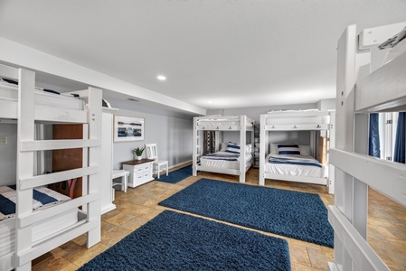 Your spacious bunk room offers plenty of comfortable sleeping space with multiple white bunk beds dressed in crisp navy-striped bedding, all set on rich hardwood floors with plush navy rugs underfoot.