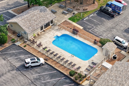 Aerial view of the property featuring a sparkling outdoor swimming pool surrounded by sun loungers, adjacent parking, and a landscaped patio area with seating and picnic tables.