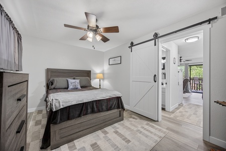 Unwind in your cozy bedroom featuring a plush bed, stylish barn door, and ceiling fan. Step through to your private balcony and enjoy serene views framed by trees.