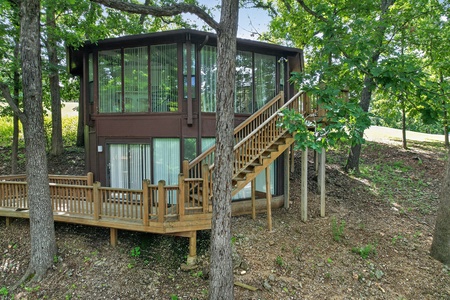 A two-story cabin nestled among mature trees in a natural woodland setting, featuring a wooden deck and staircase leading to the upper level entrance.