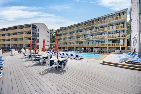 Modern property buildings frame a spacious outdoor pool deck with ample seating, umbrellas, and multiple loungers—your perfect spot to unwind under a clear sky.