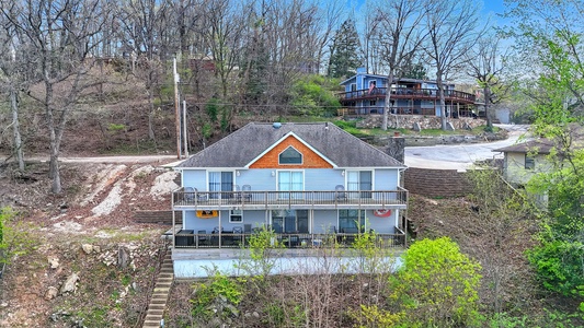 Charming lakeside cottage with multiple balconies overlooking tranquil waters, nestled among trees in a peaceful hillside setting.