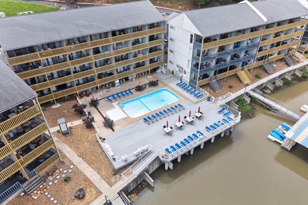 Aerial view of a waterfront resort featuring multi-story buildings with balconies, a central swimming pool, and a private dock extending over the water. The property offers direct water access and outdoor seating areas.