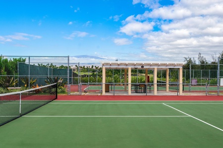 Professional tennis court with covered seating area surrounded by tropical landscaping under bright blue skies.