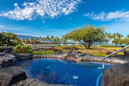 A tropical pool surrounded by volcanic rock, palm trees, and ocean-framed views under a brilliant blue sky.