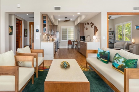 - Living area with sleek wood framed sofas and tropical accent pillows for style - Open view leads directly into a modern kitchen with warm cabinetry & bar seating - Decorative wall art and soft lighting create a relaxed and inviting coastal atmosphere
