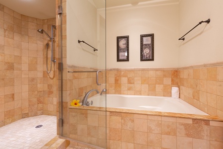 - Stylish soaking tub surrounded by natural stone tile   - Positioned beside a walk in shower for versatile bathing options   - Soft lighting and framed artwork enhance the relaxing ambiance