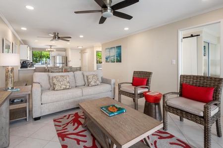 - Cozy living area with textured seating, red accents, and ceiling fans for comfort - Coastal artwork and neutral tones set the perfect tone for island relaxation - Open concept layout flows seamlessly into the kitchen and dining area