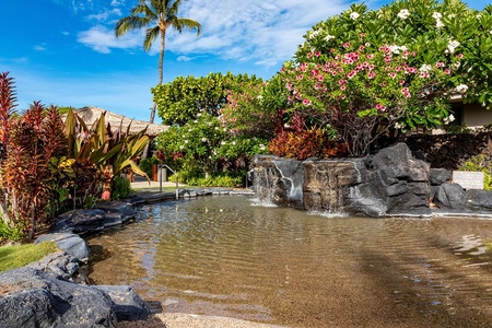 Shallow rock lined pool with cascading waterfall surrounded by vibrant tropical plants.