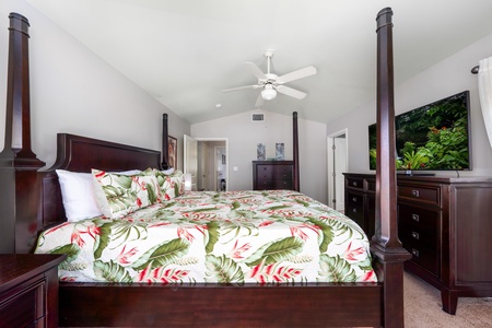 - Spacious primary bedroom with vaulted ceilings and breezy fan overhead - Tropical print bedding brings a splash of island color to the dark wood frame - View extends into the hallway with access to bathroom and closets
