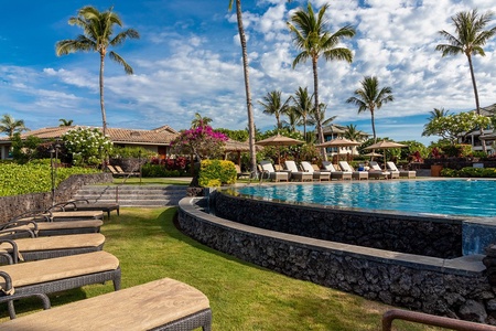 The property sits within a tropical resort setting featuring an elevated pool, manicured lawns, vibrant gardens, and swaying palms under sunny Hawaiian skies.