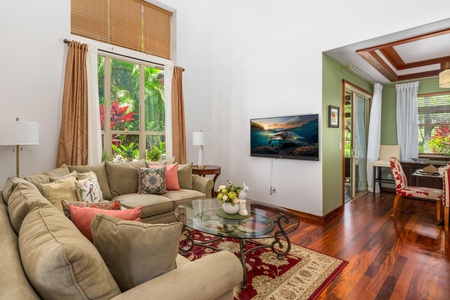 - Cozy living room with sectional sofa facing a wall mounted flat screen TV - Large windows overlook beautiful greenery for a serene tropical backdrop - Hardwood floors and warm decor add charm to this relaxing space