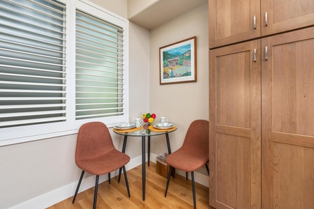 - Cozy breakfast nook with round table set for two and comfortable upholstered chairs - Bright window with shutters lets in natural light for a cheerful morning ambiance - Framed artwork adds charm while fresh fruit offers a colorful inviting touch