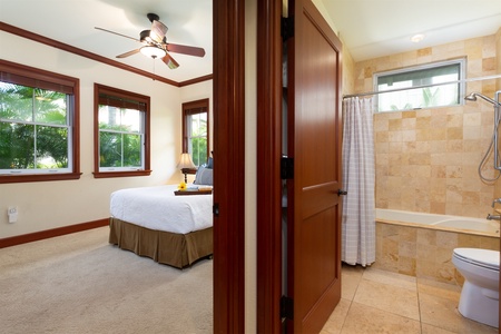 - View of bedroom and adjacent bathroom connected by a warm wood door   - Bedroom features soft carpeting, ceiling fan, and large windows with garden views   - Bathroom includes a tiled shower tub combo for versatile bathing options