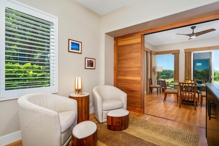 - Cozy seating nook with two cream armchairs & matching wood side tables for comfort - Sliding wood door opens to dining & living areas with views of greenery beyond - Framed wall art & soft table lighting create a warm inviting atmosphere for relaxation