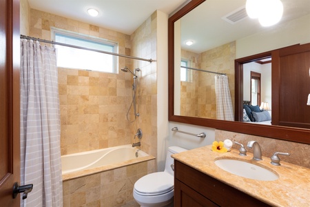 - Bright bathroom with beige stone tile and a spacious mirror above the vanity   - Tub shower combo with handheld fixture for comfort and convenience   - Elegant wood accents and fresh flower detail add a touch of style