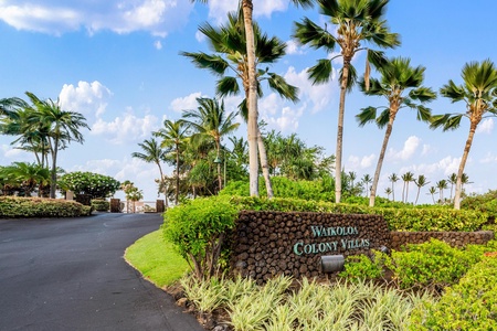 The Waikoloa Colony Villas entrance sign stands amid lush tropical landscaping, towering palms, and volcanic rock under a bright island sky.