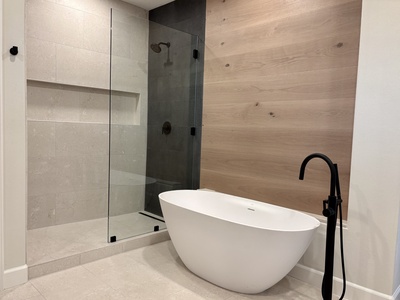 The spa-inspired primary bathroom showcases limestone floors and shower, a striking basalt accent wall, oak vanity and paneling, and a beautiful freestanding soaking tub
