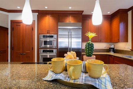 - Close up of kitchen island with granite countertop and bright pendant lighting   - Tray with yellow mugs and pastries adds a welcoming touch   - Stainless steel appliances and warm wood cabinetry create a polished look