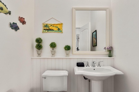 - Charming half bath with pedestal sink and elegant framed mirror above - Decorative greenery and bright wall art add a cheerful touch - Clean design with classic wainscoting creates a fresh, inviting space