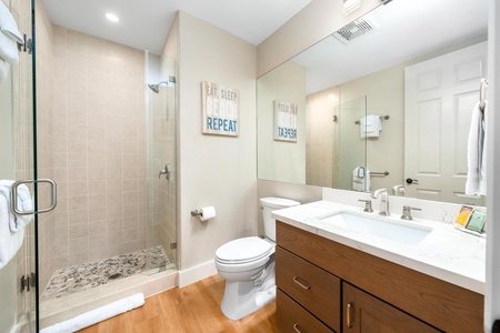 - Modern bathroom with vanity sink set in white countertop and warm wood cabinetry - Glass enclosed shower with pebble stone flooring provides a spa inspired touch - Large mirror bright lighting and decorative wall art enhance the fresh clean design