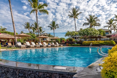 A pristine outdoor pool area framed by swaying palm trees and tropical landscaping. Enjoy lounging poolside under thatched umbrellas, with resort buildings completing this inviting Hawaiian-style retreat.
