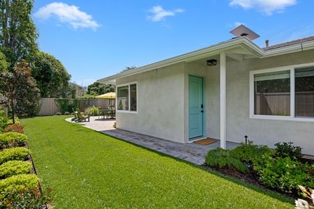 Modern single-story property featuring a charming turquoise entrance door and beautifully landscaped grounds with mature trees and outdoor seating area.