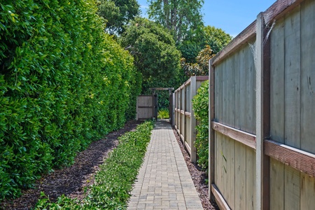 A peaceful paved walkway leads through lush greenery and wooden fencing to the property entrance.