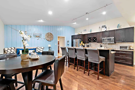 A modern kitchen with dark cabinets, a high counter with five chairs, a round dining table with four chairs, and light blue wall accents featuring a decorative clock and artwork.