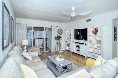 A modern living room with a sectional sofa, a coffee table, a wall-mounted TV, white shelves with decor, and large windows with sheer curtains leading to a balcony. A ceiling fan is mounted above.