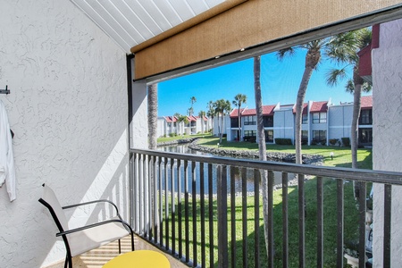 A patio with a chair and small table overlooks a canal with palm trees and apartment buildings under a blue sky.