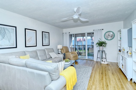 A living room with light gray sectional sofa, yellow throw blanket, framed leaf art, white shelves, wall clock, wooden floor, ceiling fan, and sliding glass doors leading to a balcony.