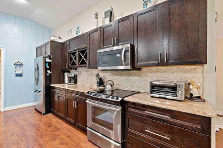 Modern kitchen with dark wood cabinets, stainless steel appliances including a refrigerator, stove, microwave, and toaster oven, granite countertops, and light-colored backsplash.