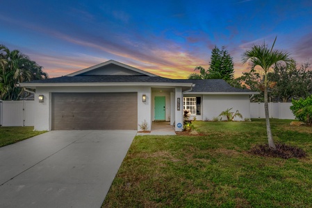 Single-story house with a two-car garage, a light blue front door, and a manicured lawn, set against a vibrant sunset sky.