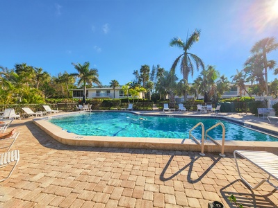 Outdoor swimming pool with surrounding lounge chairs, palm trees, and residential buildings in the background on a sunny day.