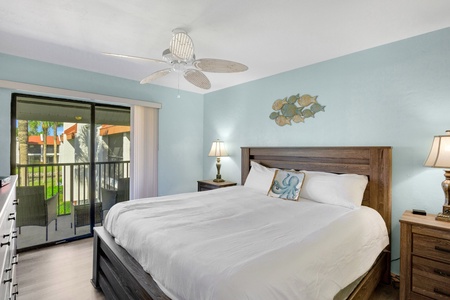 A bedroom with a large bed, two lamps on nightstands, and a sliding glass door leading to a balcony with an outside view. The room has light blue walls and a ceiling fan.