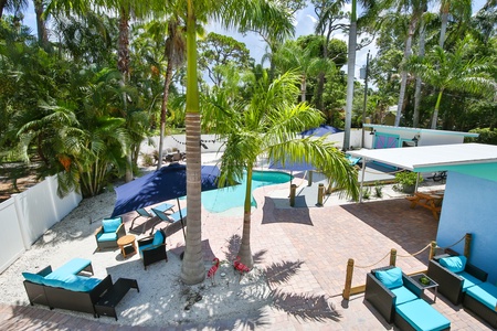 A backyard with a pool, palm trees, lounge chairs, and umbrellas. A shaded seating area and white fence are also visible.