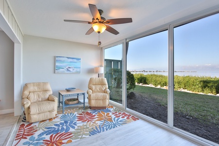 A sunroom with a ceiling fan, two beige recliners, a small table, a floral rug, and large windows offering a scenic view of greenery and a body of water.