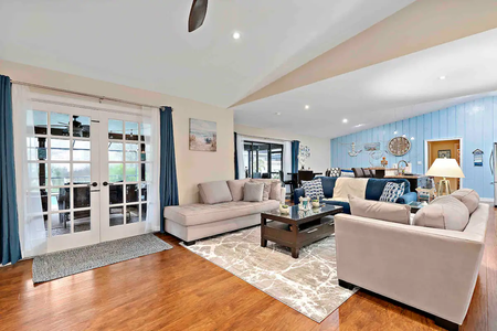 A spacious living room features light-colored sectional sofas, a wooden coffee table, hardwood floors, and French doors leading to an outdoor area. The room is accented with blue decor and wall art.