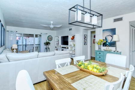 A bright, modern living and dining area with white furniture, a wooden dining table, and a tray of lemons. There is a large white sectional sofa, a wall-mounted TV, and a balcony visible in the background.