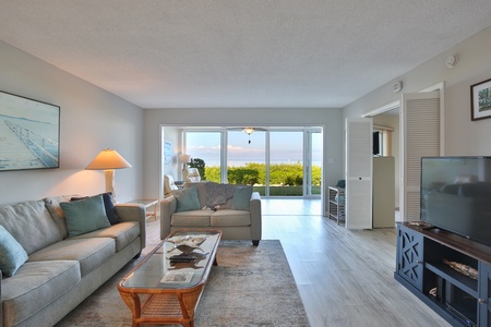 A bright living room with a beige sofa, glass coffee table, and flat-screen TV. Large sliding doors open to a patio with a view of greenery and water. An open-concept layout leads to other rooms.